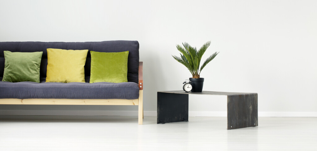Plant and clock on metal table next to a black settee with green and yellow pillow in flat interior with empty wall