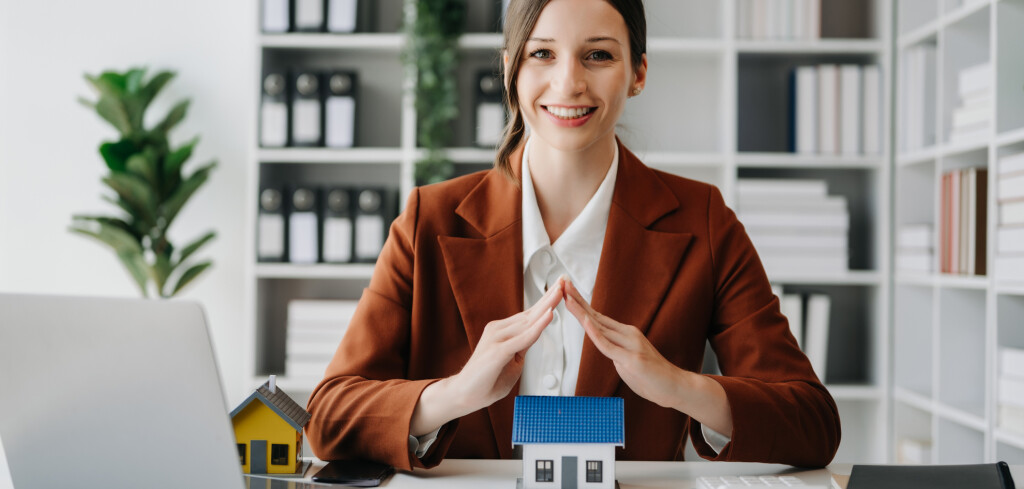 Caucasian Female real estate agent is use hands to protect red roof for the concept of real estate investment about house trading, purchase at desk in office