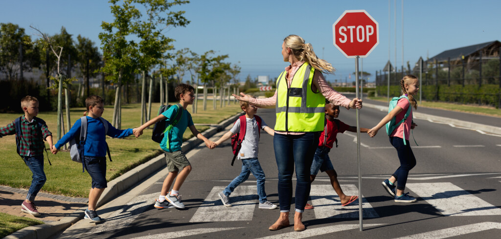 Front view of a blonde Caucasian woman wearing a high visibility vest and holding a stop sign, standing in the road and turning around on a pedestrian crossing, stopping the traffic while a diverse group of schoolchildren holding hands cross the street safely on their way to elementary school