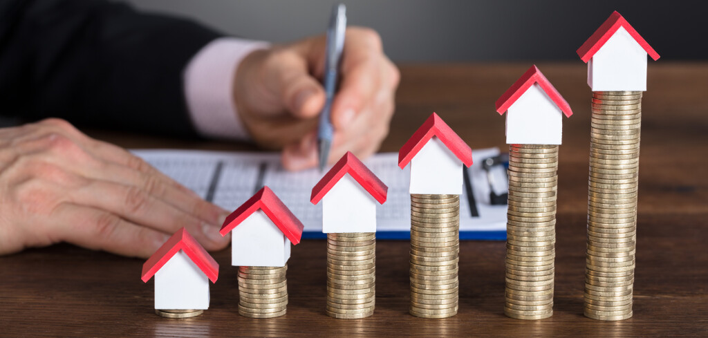 Close-up Of A Businessman Calculating Tax In Front Of Stacked Coins And House Models