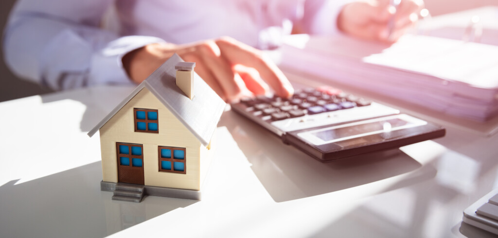 Close-up Of A Person Hand Doing Property Tax Calculation With House Model On The Table